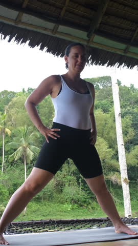 Vertical video. Woman performing yoga poses on bamboo terrace with green tropical plants and peaceful surroundings, slow motion 