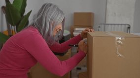 Senior woman with grey hair labeling boxes in a new home living room, signifying a move or unpacking process, surrounded by cardboard boxes and indoor plants. - Powered by Shutterstock - Get 15% off with code: PIKWIZARD15