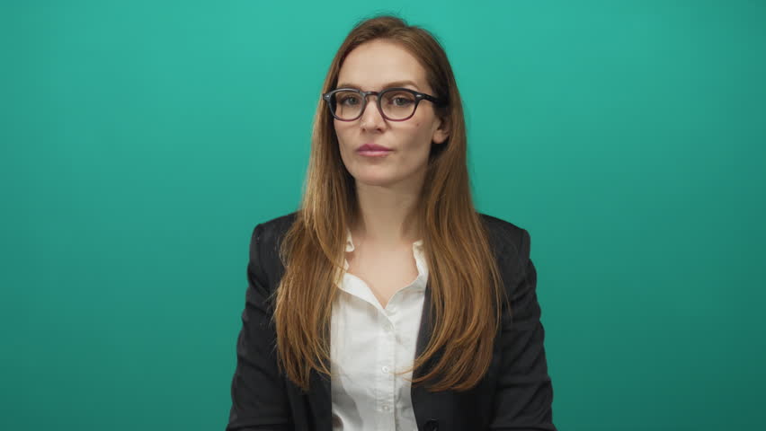Woman wearing glasses and black blazer waves hand in teal studio set, sporting white shirt and confident expression; friendly greeting.