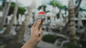 Caucasian man's hand holding a vote button outdoors in a park with blurred palm trees, highlighting democracy and civic engagement in an outdoor setting. - Powered by Shutterstock - Get 15% off with code: PIKWIZARD15