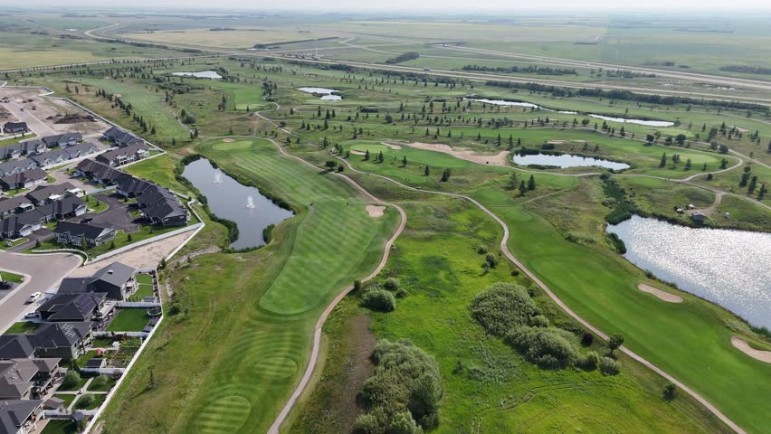Golf course with a pond and houses in the background. The golf course is surrounded by a fence and has a path leading to it