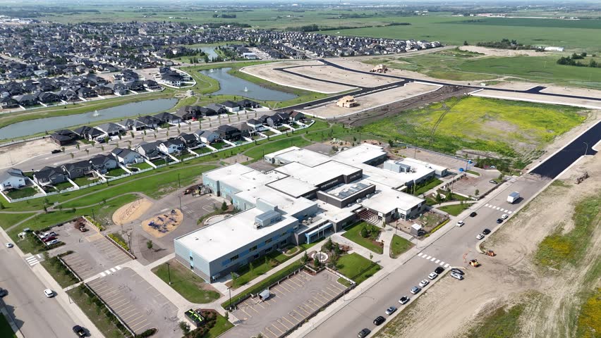 School is shown from above with a parking lot and a grassy area. The school is surrounded by houses and a large field
