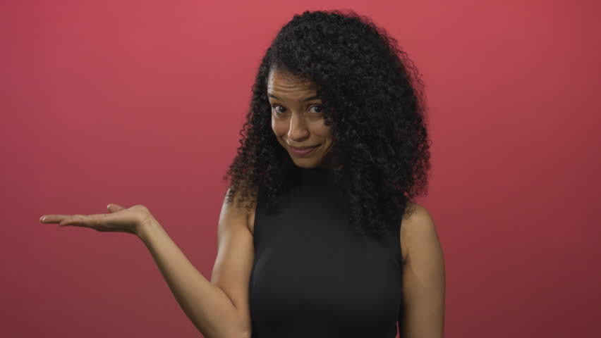 Young hispanic woman extends hand palm up and points finger at camera in red studio; warm invitation.