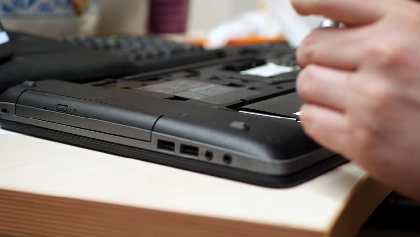 IT specialist at workshop conducts warranty diagnostic on laptop device. Screwdriver accesses compartment for maintenance. Computer repair targets upgrade. Support network ready to fix with servicing