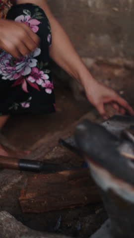 Vertical clip of An elderly Vietnamese woman adds pieces of wood to a small burning stove to keep the flames burning
