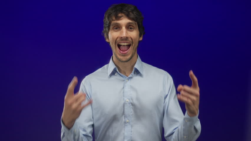 Man shows energetic rock horns with hands, sticks out tongue and grins widely in a blue studio; fun.