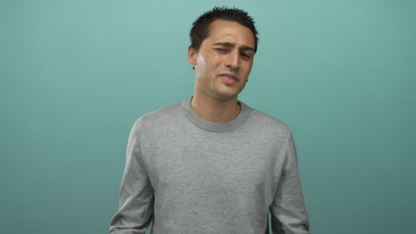 Young hispanic man wearing wristwatch holds fists to eyes in crying gesture inside blue studio set; sadness.