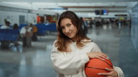Woman holding basketball in airport terminal, smiling casually indoors against busy travel backdrop, showcasing youthful, athletic, and relaxed ambiance. - Powered by Shutterstock - Get 15% off with code: PIKWIZARD15