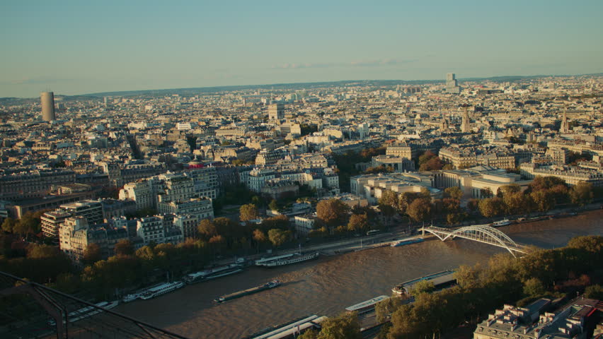 Wide aerial view of Paris in golden hour light, featuring the Seine River, Haussmannian rooftops, and a distinctive high tower in the distance.