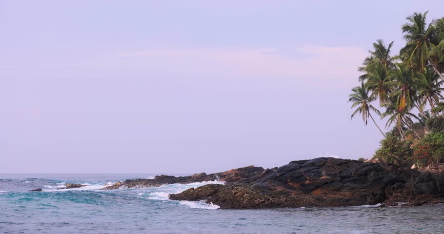 Slow motion video of waves crashing on a rocky coastline adorned with palm trees in Sri Lanka. Ideal for capturing themes of natural beauty, tropical scenery, and ocean power, this high-quality stock