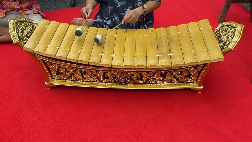 Traditional Thai musician playing wooden xylophone called Ranat Ek on ornate gold stand with red carpet background during cultural performance