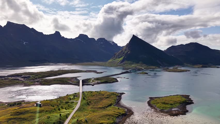 Aerial view of the Fredvang Bridge in Lofoten, Norway, surrounded by dramatic mountains, coastal water, and green summer landscape. Scenic travel destination captured by drone.