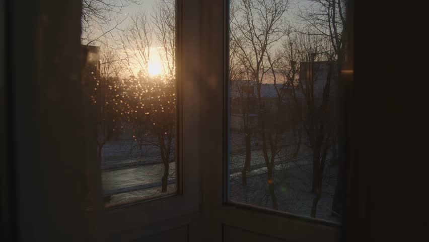 Warm sunset light streams through a window, illuminating raindrops on the glass and silhouetting bare trees and buildings outside on a quiet winter evening