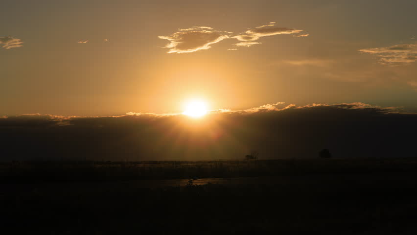 Dramatic timelapse of the sun breaking through dense storm clouds over a rural landscape, casting golden light and radiant sunbeams across the horizon. Dark cloud formations gradually give way to warm sunlight, creating a striking contrast between shadow and illumination.

The scene captures dynamic atmospheric movement with evolving cloud textures and glowing highlights, evoking themes of hope, renewal, and changing weather. Silhouetted foreground elements add depth while the sky dominates the composition.

Ideal for concepts of weather, transition, resilience, optimism, nature, climate, and environmental storytelling. Suitable for cinematic backgrounds, inspirational content, and atmospheric visual narratives.

Concepts include sunrise, sun rays, storm clouds, dramatic sky, weather change, light beams, breaking clouds, golden light, nature, landscape, hope, renewal, moody sky, and timelapse.