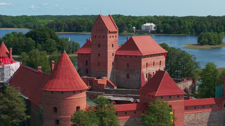 A drone view of medieval red-brick Trakai castle with multiple towers and red roofs stands on island surrounded by a lake and green trees. Lithuania