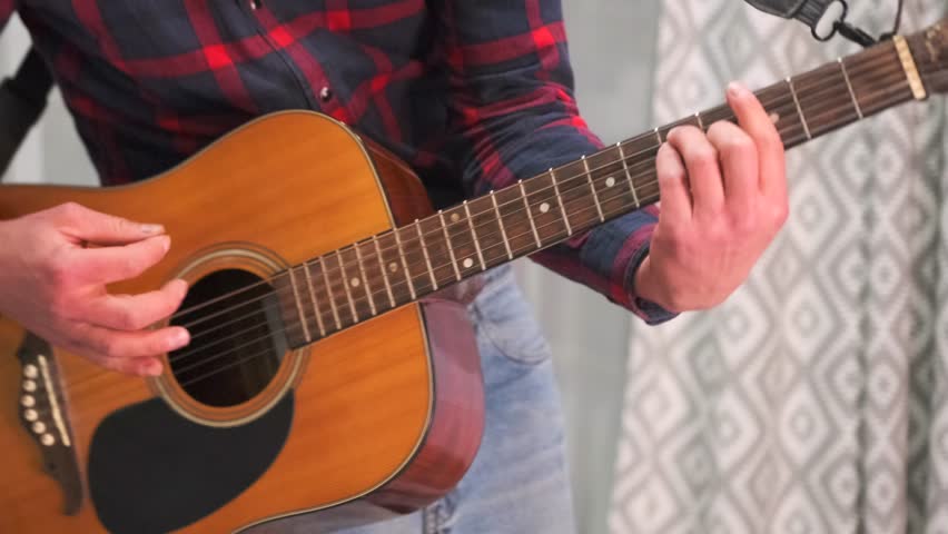 Close-up of a man in a red plaid shirt playing an acoustic guitar and gently swaying to the rhythm in a cozy indoor setting. Musical atmosphere at home, moment of personal joy and creative flow.