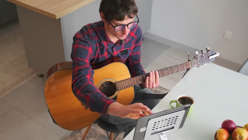 Diligent young man sits in a home kitchen with an acoustic guitar, scrolling through digital sheet music on a tablet. Focused on learning chords and melody as part of an online guitar course. 