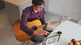 Diligent young man sits in a home kitchen with an acoustic guitar, scrolling through digital sheet music on a tablet. Focused on learning chords and melody as part of an online guitar course.  - Powered by Shutterstock - Get 15% off with code: PIKWIZARD15