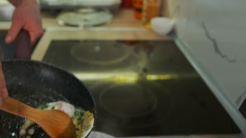 Close-up of a person using a wooden spoon to transfer freshly cooked fried eggs with green herbs from a non-stick skillet onto a decorative plate on a wooden kitchen countertop.