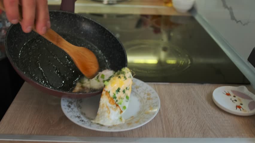 Close-up of a person using a wooden spoon to transfer freshly cooked fried eggs with green herbs from a non-stick skillet onto a decorative plate on a wooden kitchen countertop.