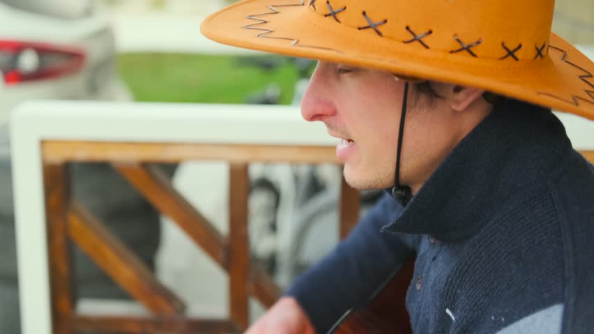 Side view of a charming young man in a traditional cowboy hat, emotionally singing and playing acoustic guitar while seated outdoors. Personal moment of musical expression. Close up.