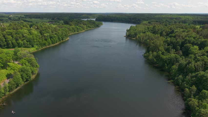 An aerial view shows wide, calm Asveja lake winding through lush green forests under a partly cloudy sky in Lithuania