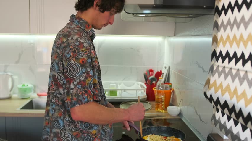 Side view man stirring noodles and eggs in pan on the stove. Homemade cooking process with simple comfort food in a cozy kitchen atmosphere.