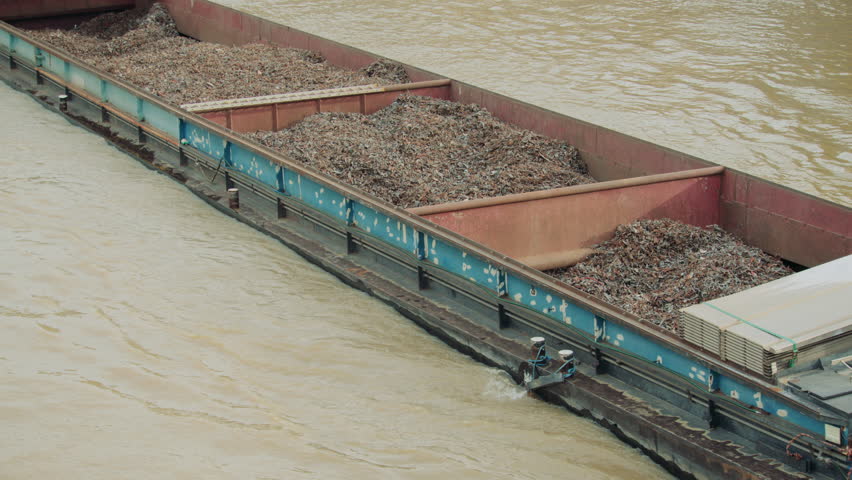 A cargo barge transports a large load of rusted scrap metal along a muddy river, highlighting industrial recycling and waterborne logistics in an urban setting.