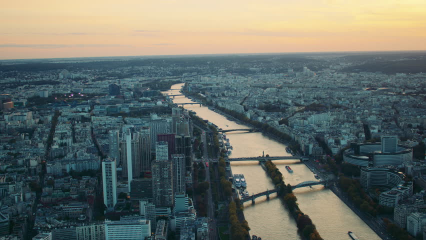 Aerial view of Paris at sunset, with the Seine River winding through the cityscape, high-rise buildings, bridges, and distant urban sprawl under a golden sky.
