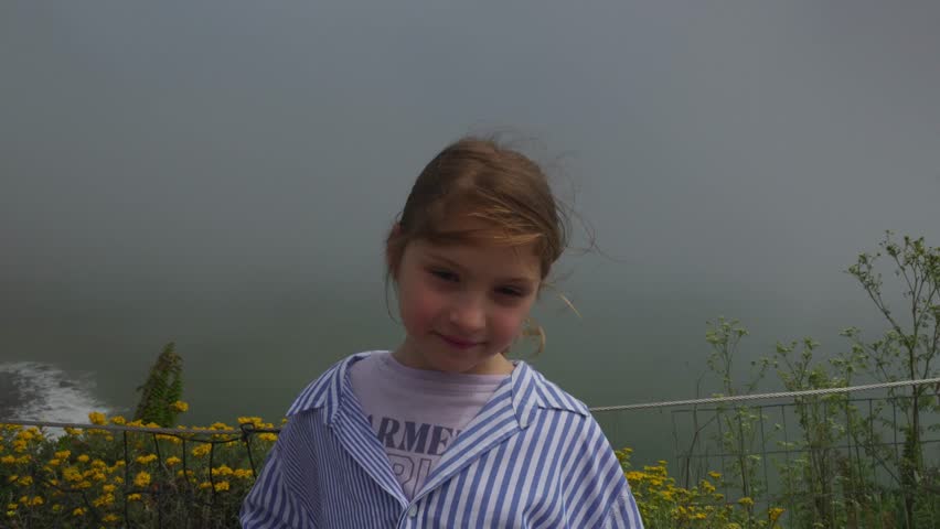 Portrait of a young girl standing by the ocean on a foggy day, with yellow wildflowers and a misty coastline in the background.
