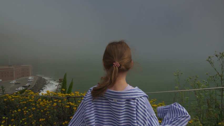 Young girl with ponytail standing near yellow wildflowers, looking at foggy coastline and Fort Point by the sea.
