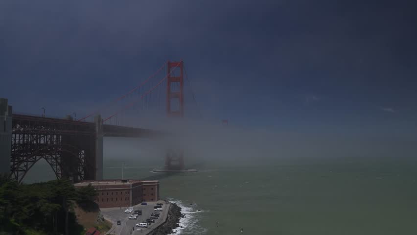 Golden Gate Bridge emerging from thick fog above Fort Point and the San Francisco Bay, seen from an elevated viewpoint.
