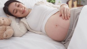 Asian young pregnant woman gently stroking belly while lying down on bed. Attractive expectant mother looks at her baby bump with happiness and love, embracing beauty of pregnancy in bedroom at home. - Powered by Shutterstock - Get 15% off with code: PIKWIZARD15