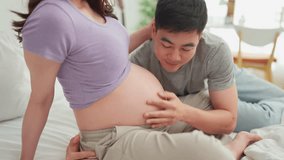 Asian husband and pregnant wife sit together on cozy bed in bedroom. Attractive expectant couple gently touching and listening to baby bump, sharing loving moment ready to become parents in future. - Powered by Shutterstock - Get 15% off with code: PIKWIZARD15