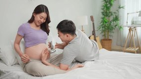 Asian expectant couple sharing loving moment, ready to become parents. Attractive pregnant wife and husband placing headphones on belly to play music for baby while sitting on cozy bed in bedroom. - Powered by Shutterstock - Get 15% off with code: PIKWIZARD15