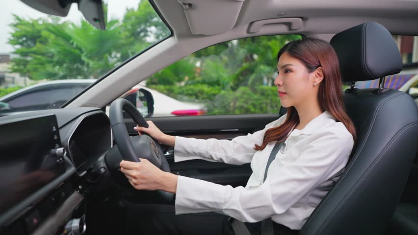 Beautiful Asian woman fastening her seatbelt while driving a car. Attractve young woman employee sit inside the vehicle, feel happy and enjoy showing safe and protected driving while driving to work.