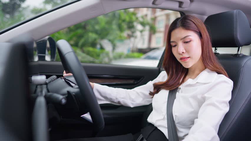 Beautiful Asian woman fastening her seatbelt while driving a car. Attractve young woman employee sit inside the vehicle, feel happy and enjoy showing safe and protected driving while driving to work.