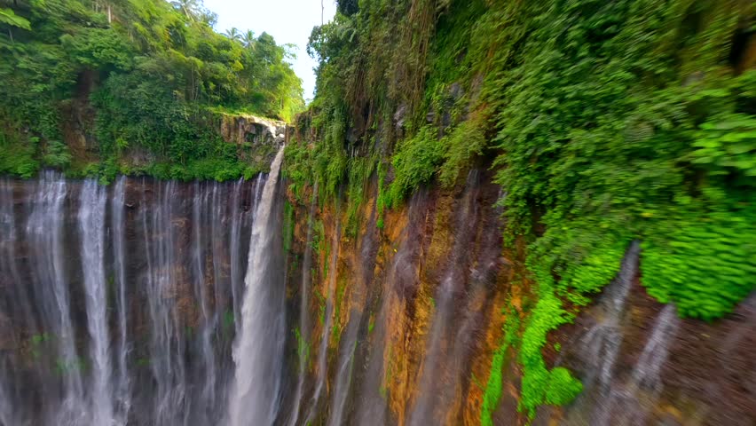 Cinematic FPV drone shot diving through the stunning multi-stream Tumpak Sewu waterfall, surrounded by lush rainforest cliffs in East Java, Indonesia.