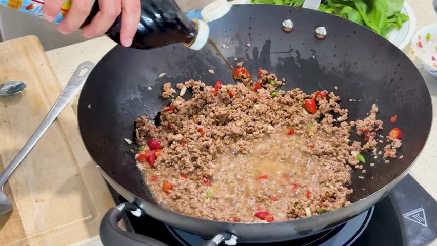 Hand pours fish sauce from a bottle into a wok with boiling ground meat and chopped vegetables, under bright kitchen lighting, top-down view
