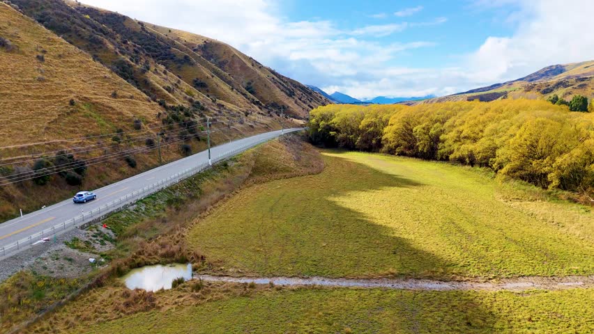 Drone footage glides above a winding highway through golden autumn trees and hills near Wanaka, New Zealand, under bright natural daylight