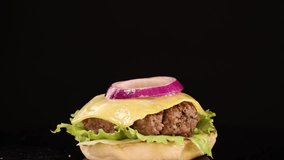 A sesame seed bun top falls onto a stacked beef burger with lettuce, cheese, tomato, and onion, shot in slow motion against a black background with studio lighting - Powered by Shutterstock - Get 15% off with code: PIKWIZARD15