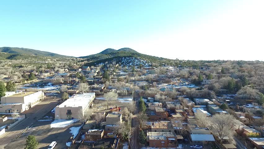 Aerial drone footage flying over a residential neighborhood in Santa Fe, New Mexico during winter. Snow patches are visible on rooftops and the ground, contrasting with distant green rolling hills.
