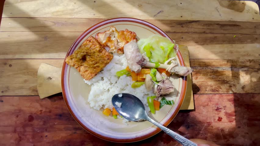 someone is spooning rice with warm soup served with side dishes of tempeh and chicken in a brown ceramic plate on an old wooden table, chicken bone soup with various vegetables