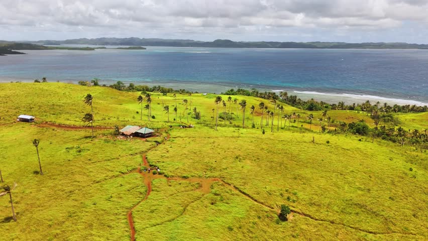Aerial view of Corregidor Island, Siargao Islands, Philippines shows grassy terrain, palm trees, coastal huts, Pacific Ocean, and surrounding islets under bright daylight in tropical conditions.
