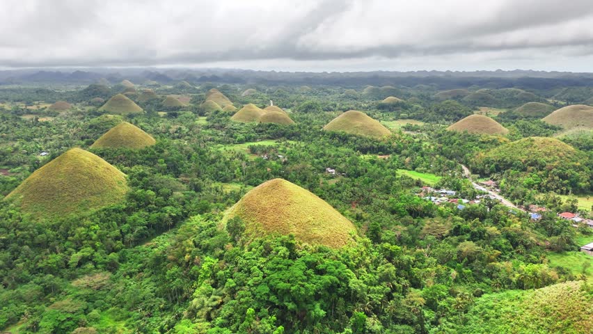 Drone reveals the famed Chocolate Hills in Bohol, Philippines—symmetrical grass-covered mounds spread across a lush tropical landscape, with distant villages and cloudy skies