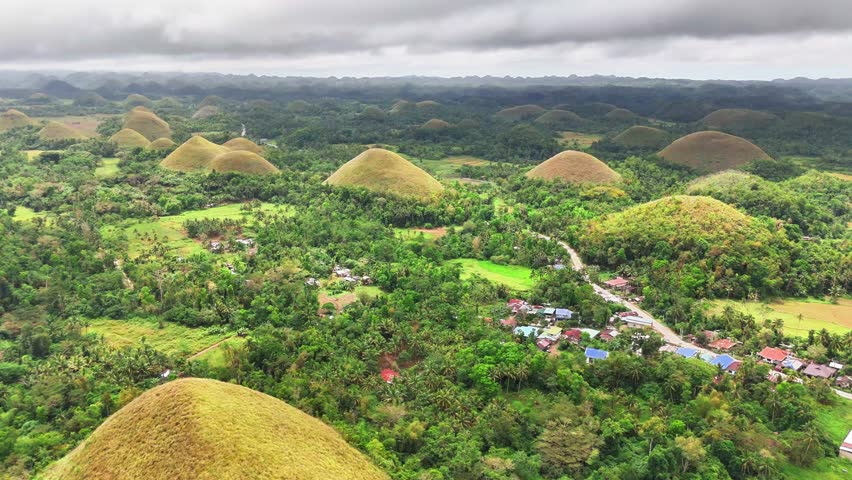 Stunning aerial pull out shot reveals the iconic Chocolate Hills of Bohol, Philippines, scattered across lush tropical forest and villages under dramatic overcast skies on a humid day.