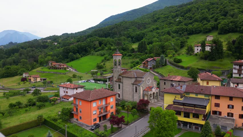 Drone shot of a stone church surrounded by colorful homes and green hills in the Alpine foothills. Shot at Villa di Tirano, Sondrio, Italy (Italia)