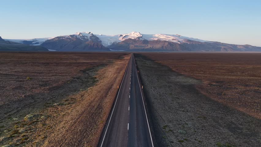 Aerial view of the stark, straight road cutting through the barren landscape towards distant snowy mountains under a clear sky, Iceland roads, Iceland.
