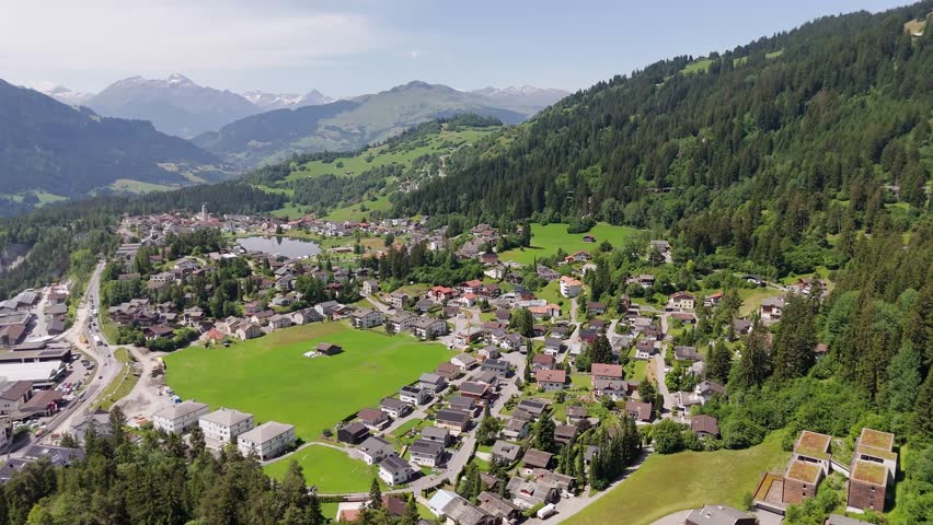 Aerial view of Swiss town with neighborhood , green pasture and cars on street. Wide shot. Sunny day in summer. Alps mountains in distance.