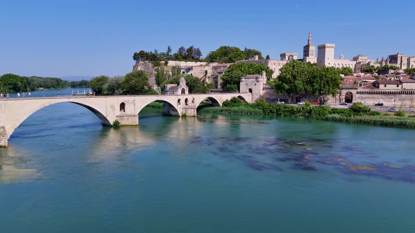 A wide zoom-out shot reveals the historic Pont d’Avignon spanning the Rhône River, with the impressive Palais des Papes rising behind it, showcasing Avignon’s iconic medieval skyline.